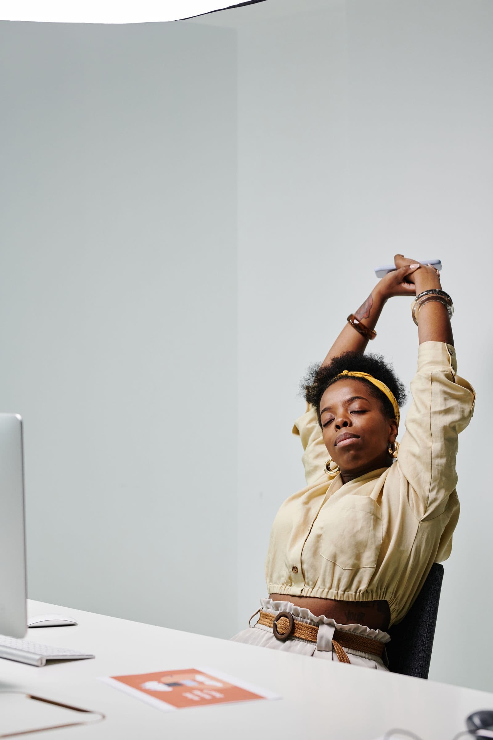A woman at an office desk stretching both arms overhead with her eyes closed, taking a brief break