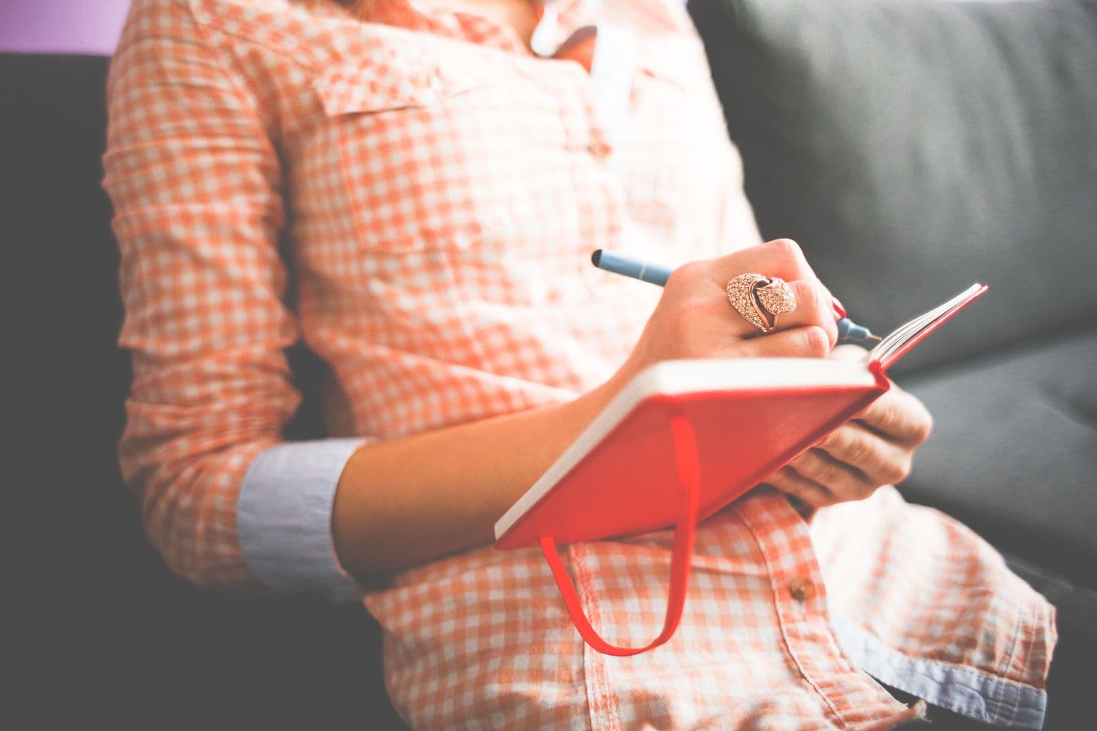 A close-up of a woman writing in a red journal with a pen while sitting on a sofa