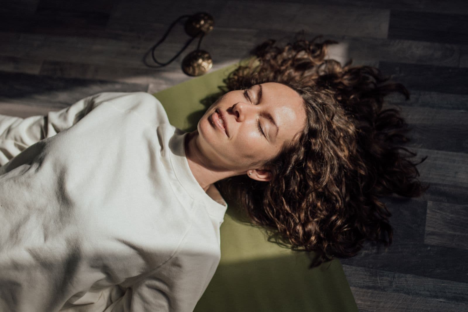 A woman lying on a green yoga mat with her eyes closed, relaxed, with meditation chimes resting on the floor beside her