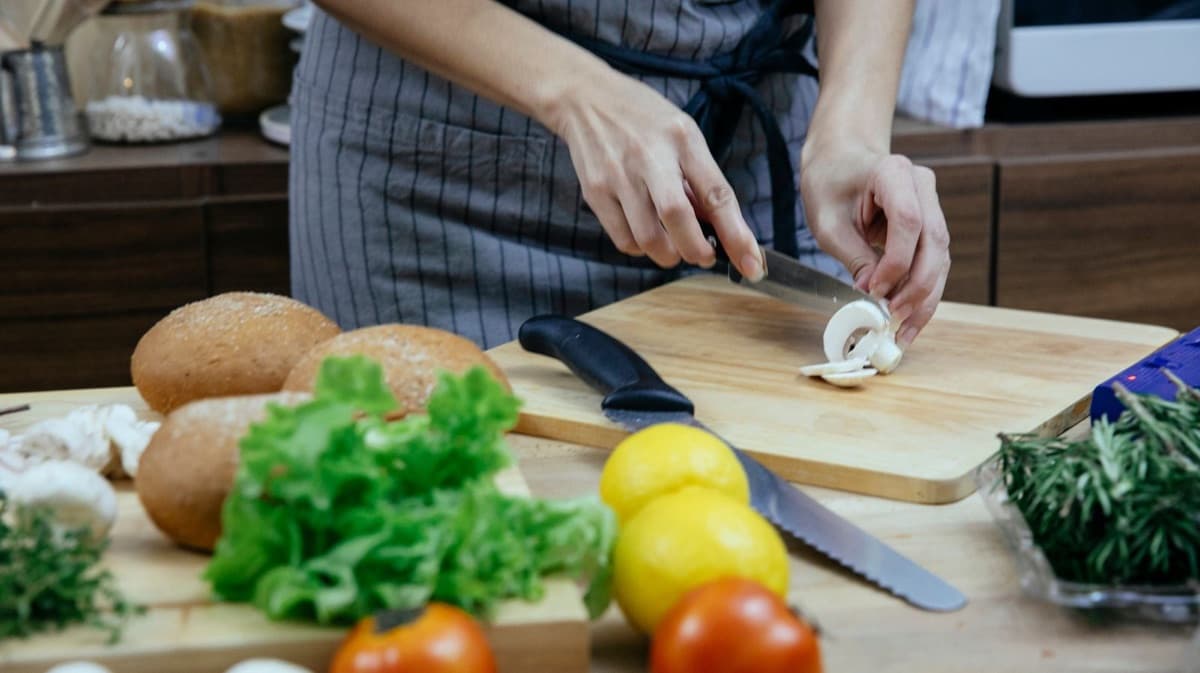 A person in an apron slicing fresh mushrooms on a wooden cutting board, surrounded by tomatoes, lettuce, lemons and herbs