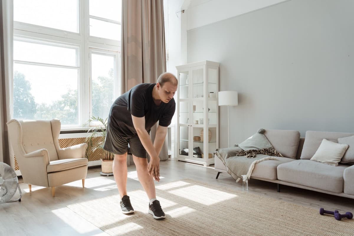 A man in a black t-shirt doing a standing side stretch in a bright, sunlit home living room, with a pair of dumbbells visible on the rug